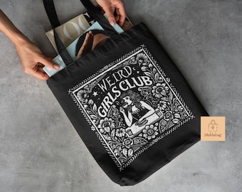 May include: Black tote bag with the words "WEIRD GIRLS CLUB" in a white floral design. The bag is filled with magazines and held by hands. A small, square, tan-colored tag with a logo is in the lower right corner.