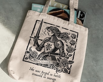 Pode incluir: Saco tote bege com uma ilustração a preto e branco de uma cavaleira segurando uma espada, rodeada por rosas. O texto diz "Main Character Energy" e "She was forged in battle, crowned in courage."