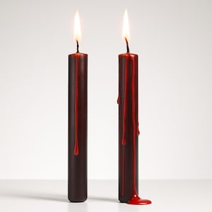 May include: Two lit, dark brown candles with red wax dripping down the sides. The candles have a cylindrical shape and are set against a white background. The flames are bright and yellow.