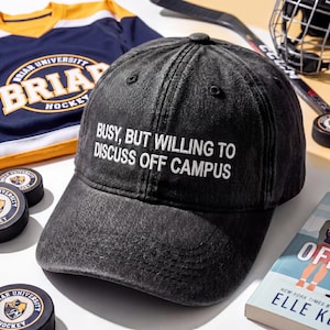 May include: A black baseball cap with the text "BUSY, BUT WILLING TO DISCUSS OFF CAMPUS" in white. The cap is part of a collection of hockey-related items, including a jersey, pucks, and a hockey helmet.