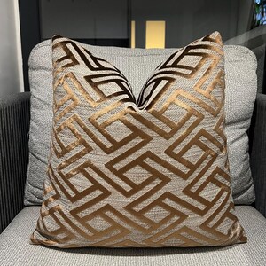 Puede incluir: Un cojín cuadrado decorativo con un patrón geométrico en tonos marrón y dorado. El cojín está colocado sobre una silla gris tapizada, mostrando su tejido texturizado y su diseño. El patrón es una serie repetida de formas angulares.