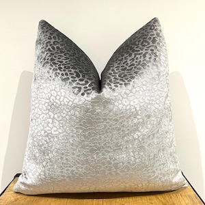 Puede incluir: Un cojín cuadrado de tela de terciopelo plateado metálico con un patrón de leopardo texturizado en relieve. El cojín tiene una parte superior central distintivamente pellizcada, creando una forma estructurada con un acabado brillante.