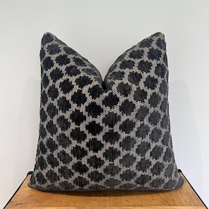 May include: A decorative throw pillow with a dark gray and beige geometric pattern. The pillow is square-shaped and has a textured surface, adding a touch of elegance to any living space. The pillow is placed on a wooden surface.