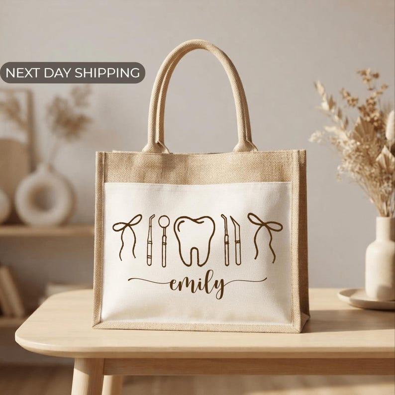 A beige tote bag with a dental-themed design printed on it, featuring a tooth and dental tools, and the name "emily" written below. The bag is placed on a wooden surface, and there is a vase of dried flowers in the background.
