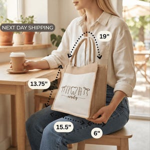 A woman sitting at a table with a coffee cup in front of her, holding a large tote bag with a printed design.