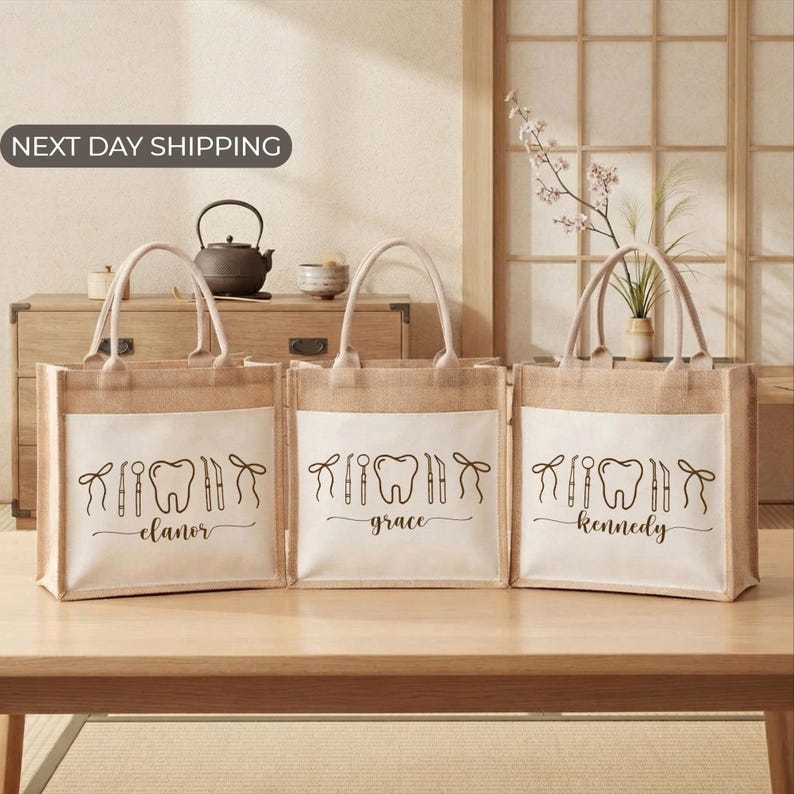 Three beige tote bags with the same design printed on them, each containing a different name: Elanor, Grace, and Kennedy. The bags are placed on a wooden surface, and there is a vase with flowers in the background.