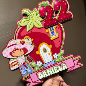 May include: A colorful strawberry-themed birthday cake topper featuring a cartoon girl holding a cake, a strawberry house, and the number 22. The name "DANIELA" is displayed on a pink banner. The design is predominantly red, pink, and green.