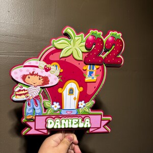 May include: A colorful strawberry-shaped birthday decoration featuring a cartoon character, a house, and the number 22. The name "DANIELA" is displayed on a pink banner. The decoration is held by a clear stick.