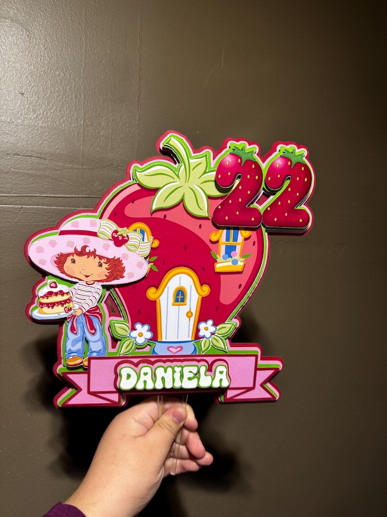 May include: A colorful strawberry-shaped birthday decoration featuring a cartoon character, a house, and the number 22. The name "DANIELA" is displayed on a pink banner. The decoration is held by a clear stick.