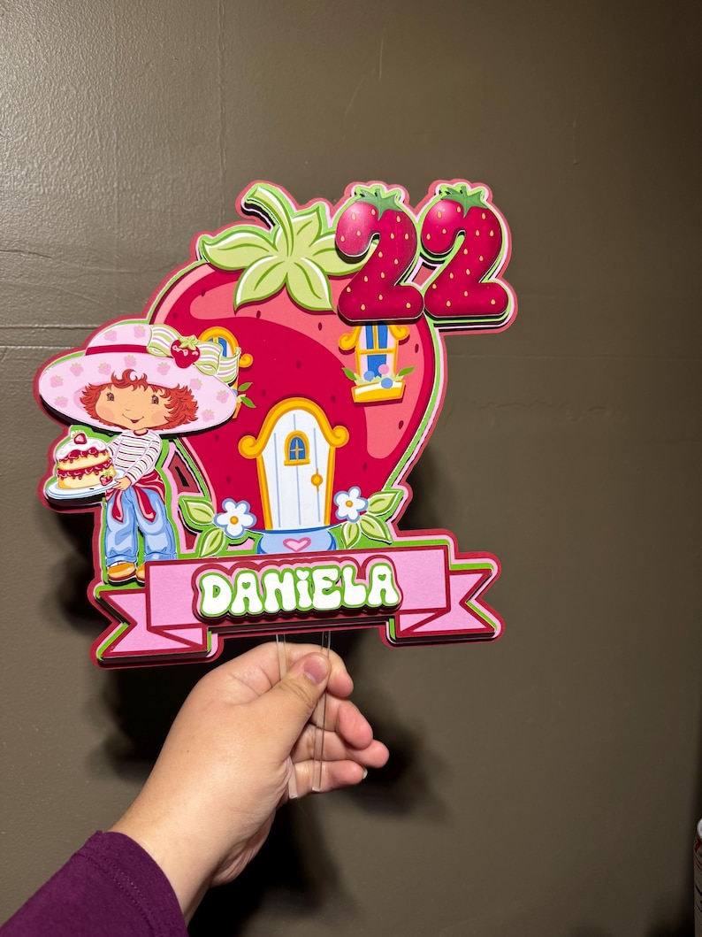 May include: A colorful, layered decoration featuring a strawberry-shaped house with a door and window. A cartoon character in a pink hat and a banner that says "DANIELA" is included. The number "22" is at the top. The decoration is held by a hand.
