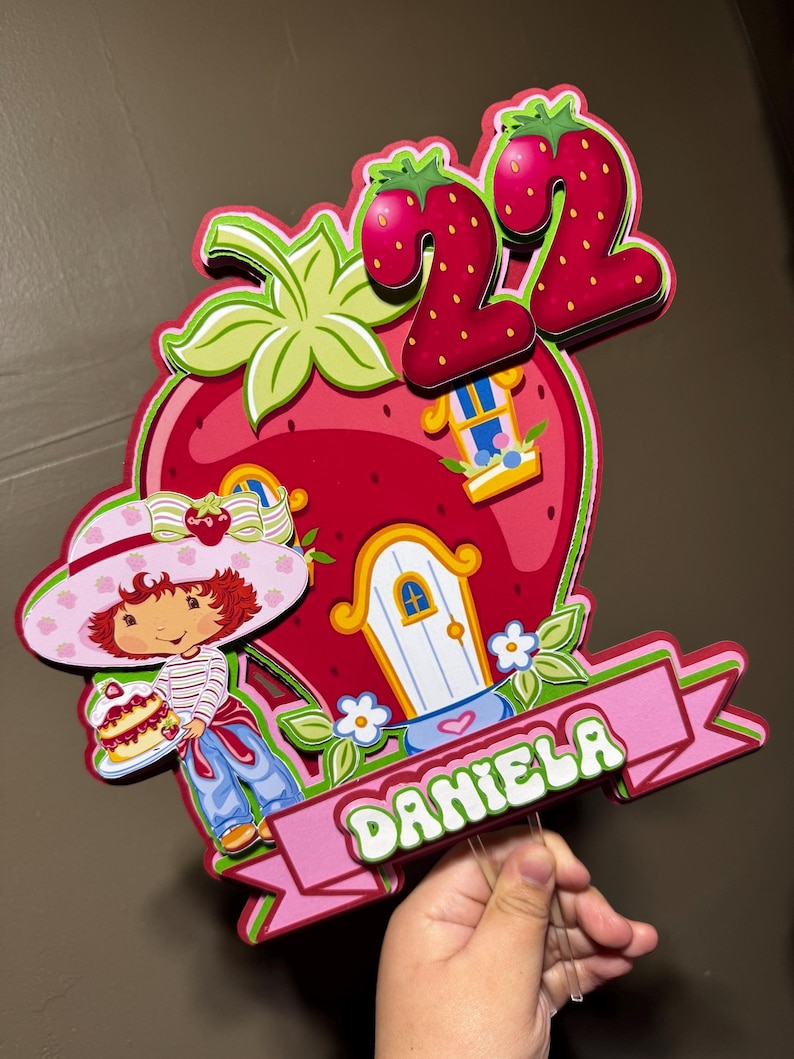 May include: A colorful strawberry-themed birthday cake topper featuring a cartoon girl holding a cake, a strawberry house, and the number 22. The name "DANIELA" is displayed on a pink banner. The design is predominantly red, pink, and green.