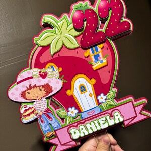 May include: A colorful strawberry-themed birthday cake topper featuring a cartoon girl holding a cake, a strawberry house, and the number 22. The name "DANIELA" is displayed on a pink banner. The design includes red, pink, green, and blue elements.