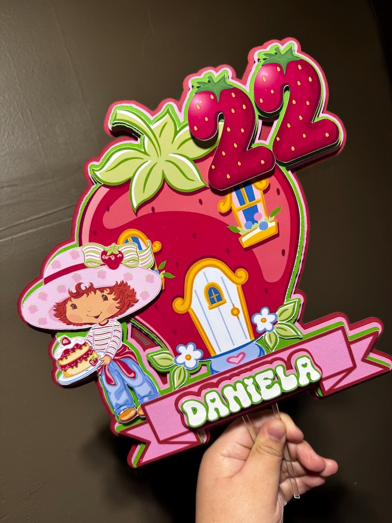 May include: A colorful strawberry-themed birthday cake topper featuring a cartoon girl holding a cake, a strawberry house, and the number 22. The name "DANIELA" is displayed on a pink banner. The design includes red, pink, green, and blue elements.