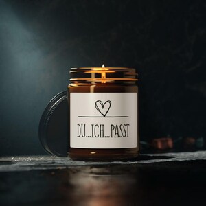 Könnte beinhalten: Eine brennende Kerze in einem braunen Glas mit goldenem Deckel. Das Glas hat ein weißes Etikett mit einem handgezeichneten Herz und dem deutschen Text "DU...ICH...PASST" in Schwarz. Der Deckel liegt daneben.