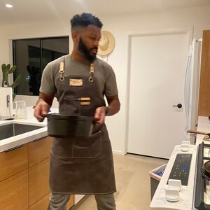 May include: A person wearing a gray t-shirt and a brown apron with leather straps is holding a black cast iron pot in a kitchen.