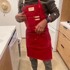 May include: A red canvas apron with adjustable straps and a leather patch that says "Genuine Tommy." The apron has two large pockets and a smaller pocket on the front.