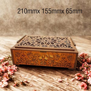 May include: A decorative wooden box with intricate floral cutouts on the lid and detailed carvings on the front. The box measures 210mm x 155mm x 65mm. Pink flowers are scattered around the box on a wooden surface.