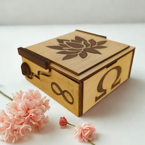 Puede incluir: Caja de madera de color marrón claro con un diseño de flor de loto en la tapa. Los lados están grabados con el símbolo del infinito y la letra griega omega. La caja tiene detalles en marrón oscuro y un cierre de cierre.