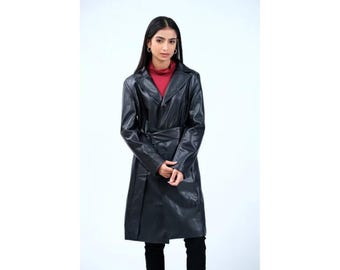 Pode incluir: Um trench coat longo de couro preto com cinto na cintura e gola entalhada. O casaco é usado sobre uma gola alta vermelha. A modelo está usando calças pretas. O casaco parece ser na altura do joelho.
