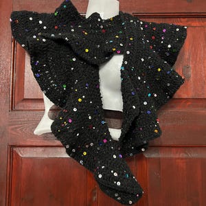 May include: A black knitted scarf adorned with colourful sequins. The scarf is displayed on a white mannequin. The sequins are in various colours, including silver, gold, red, and blue. The scarf has a textured appearance and is likely handmade.