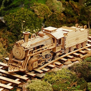 May include: A detailed wooden model of a steam train, featuring intricate laser-cut designs. The train is light brown and sits on a wooden track, surrounded by green moss and foliage. The model includes a locomotive, tender, and passenger car.