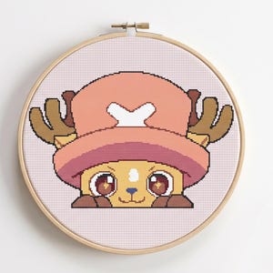 May include: A cross-stitch embroidery of a cartoon character in a wooden hoop. The character has a pink hat with a white cross, brown antlers, and large brown eyes. The background is a light pink grid.