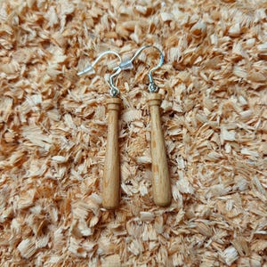 Könnte beinhalten: Ein Paar Baseballschläger-Ohrringe aus Holz mit silberfarbenen Haken. Die Ohrringe sind hellbraun und haben ein detailliertes, miniaturisiertes Schlägerdesign. Der Hintergrund ist ein Bett aus hellbraunen Holzspänen.