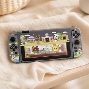 Op de afbeelding: Een grijze handheld spelcomputer met een decoratieve skin met Animal Crossing-personages zoals Tom Nook, Isabelle en K.K. Slider. De console toont een scherm met een dorpsscène met diverse gebouwen en geanimeerde dierenfiguren.