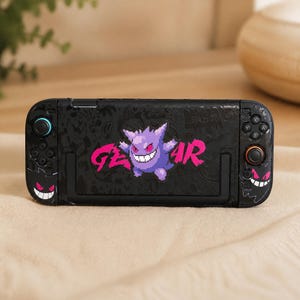 Op de afbeelding: Een zwarte draagbare spelcomputer met een gepixelde paarse Gengar-figuur en de tekst GENGAR in felroze. Het apparaat bevat zwarte controllers met geïntegreerde knoppen, een joystick en extra afbeeldingen van het gezicht van Gengar.