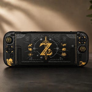 May include: A black handheld gaming console featuring gold-colored graphic designs. The center displays a large gold sword emblem with a stylized Z, surrounded by smaller gold icons, symbols, and intricate line work on a dark matte surface.