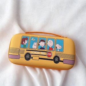 Op de afbeelding: Gele rechthoekige etui met een schoolbusontwerp en stripfiguren. De etui heeft een handvat en een ritssluiting. De schoolbus heeft zwarte wielen, een stopbord en de tekst "SCHOOL BUS".