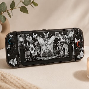 Puede incluir: Una consola de juegos portátil negra con un estampado gráfico blanco detallado de una mariposa central grande, mariposas más pequeñas, marcos de fotos ornamentados, relojes y candelabros. El dispositivo tiene un joystick azul y uno rojo.