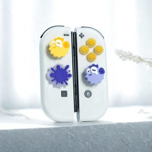 Könnte beinhalten: Weißer Gamecontroller mit bunten dekorativen Knöpfen. Die Knöpfe zeigen gelbe und lila Cartoonfiguren und ein blaues Tintenspritzer-Design. Der Controller ist geöffnet und zeigt die internen Komponenten.