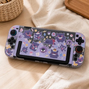 May include: A handheld gaming console featuring a light purple skin decorated with various purple Gengar character stickers, small hearts, stars, cherries, and Poké Ball icons. The device includes black joysticks, directional buttons, and action buttons.