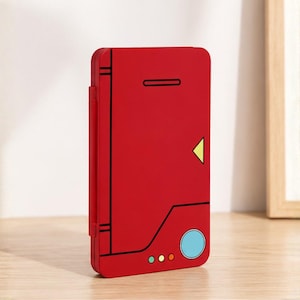 Puede incluir: Un estuche rectangular rojo con un lado articulado, que presenta detalles de líneas negras, un acento triangular amarillo, un círculo grande azul claro y tres círculos pequeños en verde, amarillo y rojo en el borde inferior frontal.