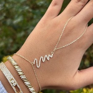 Könnte beinhalten: Eine Sammlung goldfarbener Schmuckstücke. Das Bild zeigt eine Hand, die mit einer zarten Goldhalskette im Herzschlag-Design und drei goldenen Armbändern geschmückt ist. Die Armbänder sind mit funkelnden, klaren Steinen verziert.