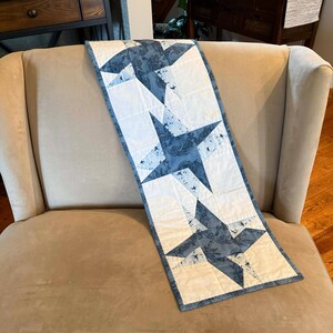 May include: A rectangular table runner featuring a star pattern. The stars are a mix of white and blue fabric with a floral design. The runner is placed on a beige armchair.