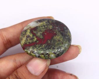 Puede incluir: Una piedra preciosa redonda y pulida con una mezcla de colores. La piedra presenta una base verde oliva y amarilla con llamativas vetas rojas. La piedra se sostiene en una mano.