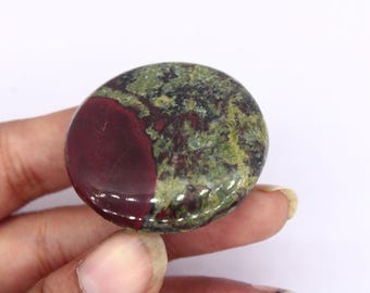 Puede incluir: Una piedra redonda y pulida con una mezcla de colores. La piedra presenta una sección burdeos oscuro a la izquierda y un patrón moteado verde y negro a la derecha. La piedra se sostiene en una mano sobre un fondo blanco.