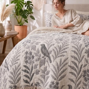 May include: A cream-colored quilted bedspread featuring a repeating dark gray botanical pattern of birds perched on leafy branches and floral clusters. The edges of the textile are finished with a decorative fringe trim along the bottom border.