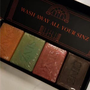 May include: A black rectangular box containing four rectangular soap bars in orange, green, pink, and dark brown. Each bar is wrapped in clear plastic and embossed with the word SINZ. The box lid features the text WASH AWAY ALL YOUR SINZ.