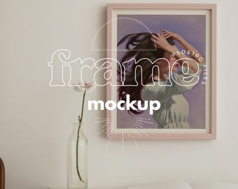 Op de afbeelding: Een ingelijste kunstafdruk-mockup met een vrouw met wapperend haar, gekleed in een lichtgroene jurk, tegen een paarse achtergrond. De lijst is zachtroze. Een heldere glazen vaas met een bloem en een open boek staan op een witte tafel. Tekst op de afbeelding luidt "frame mockup".