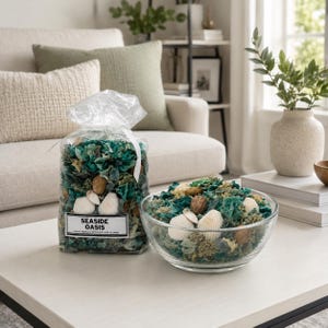 May include: A clear glass bowl and a clear plastic bag filled with decorative potpourri. The potpourri includes seashells, nuts, and teal, green, and beige dried botanicals. The bag has a label that reads "Seaside Oasis".