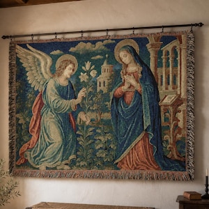 Horizontal woven blanket wall hanging featuring Archangel Gabriel kneeling before the Virgin Mary in an Annunciation scene, with lily flowers, halos, soft robes, garden landscape, old architecture, muted vintage colors, and fringed textile edges.