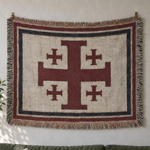 May include: A rectangular woven throw blanket featuring a large dark red Jerusalem cross centred on a light beige background. The design is framed by a dark blue border and a thin red border, with decorative fringed edges on all four sides.