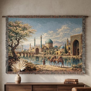 May include: Woven tapestry depicting an Eastern cityscape with a river, bridge, and domed buildings. The artwork features camels, a tree with pink blossoms, and a serene landscape. The tapestry is framed with a brown border and fringe.