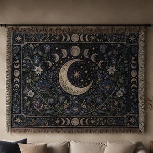 May include: A rectangular woven wall tapestry featuring a dark blue background with a central crescent moon, a star, and a surrounding lunar cycle. The design includes intricate floral patterns in shades of blue, green, and cream with fringed edges.