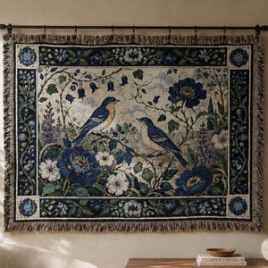 May include: A rectangular woven wall tapestry featuring two blue birds perched on branches amidst large dark blue flowers and green foliage. The design is framed by a decorative border of blue blossoms and leaves, finished with a fringed edge.