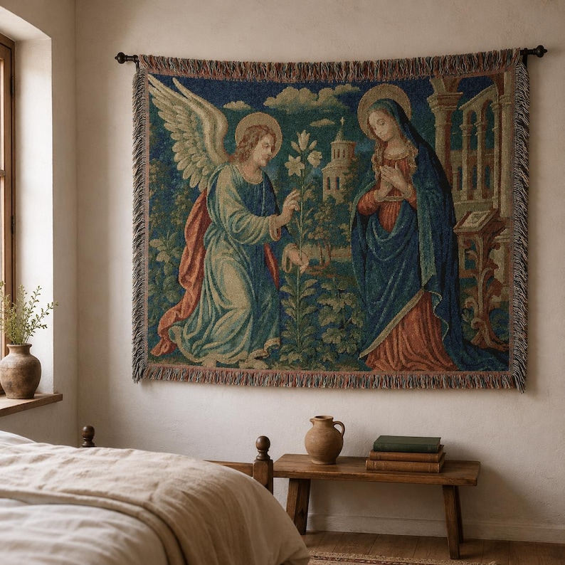 Horizontal woven blanket wall hanging featuring Archangel Gabriel kneeling before the Virgin Mary in an Annunciation scene, with lily flowers, halos, soft robes, garden landscape, old architecture, muted vintage colors, and fringed textile edges.