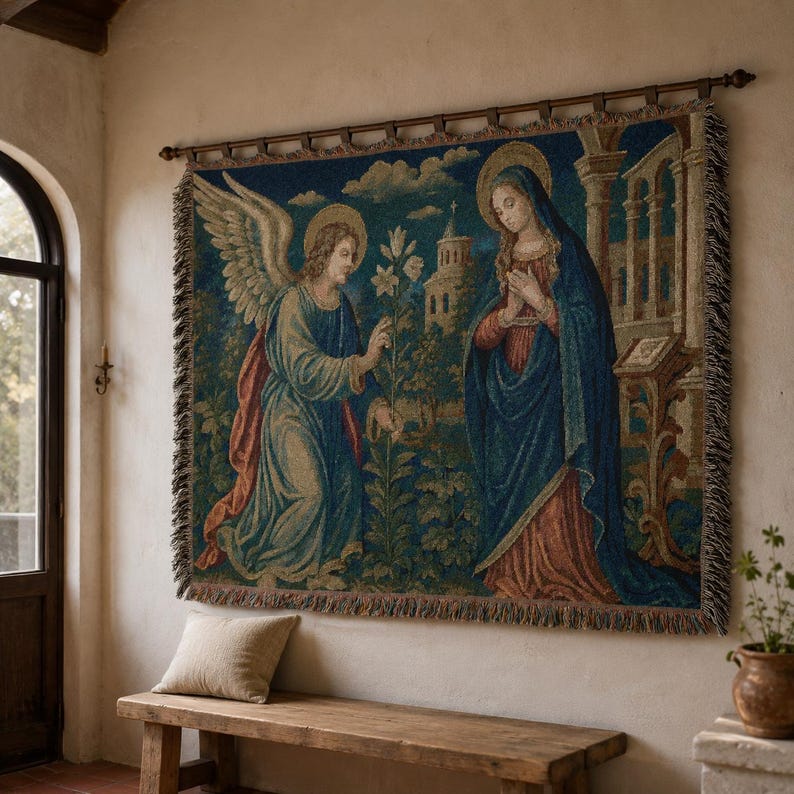 Horizontal woven blanket wall hanging featuring Archangel Gabriel kneeling before the Virgin Mary in an Annunciation scene, with lily flowers, halos, soft robes, garden landscape, old architecture, muted vintage colors, and fringed textile edges.